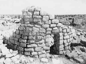 A black and white photo of the ruins with large stone bricks making up some walls and a door on the right side. 