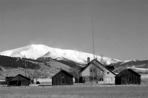 A black and white photo of the ranch complex with buildings before mountains in the background in black and white.