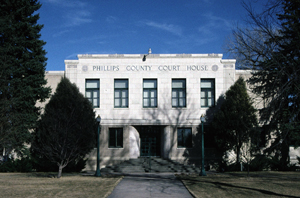 A view of the courthouse from the front with path leading up to a white building with falt roof and evenly spaced windows on the second floor. On either side of the picture are two sets of trees.