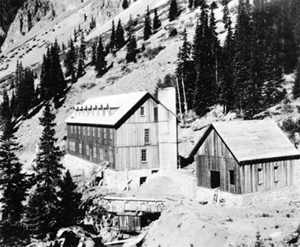A black and white photo of two of the buildings with gabled roofs and the one on the left with dormers and chimney. There is a very large slope in the background ascending to the right and sparsely covered with trees.