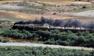 A picture of the train moving to the left with a black smoke trail coming from the front with trees and plains landscapes on before and behind it.