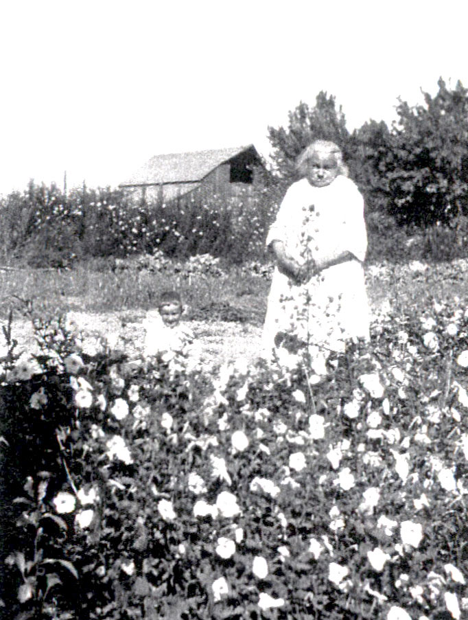 An old woman and small boy amongst flowers in an historic photo of the Koch Farm.  The original milk barn is in the background.