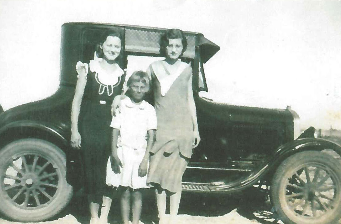 Historic image showing two women and a girl standing in front of a car.