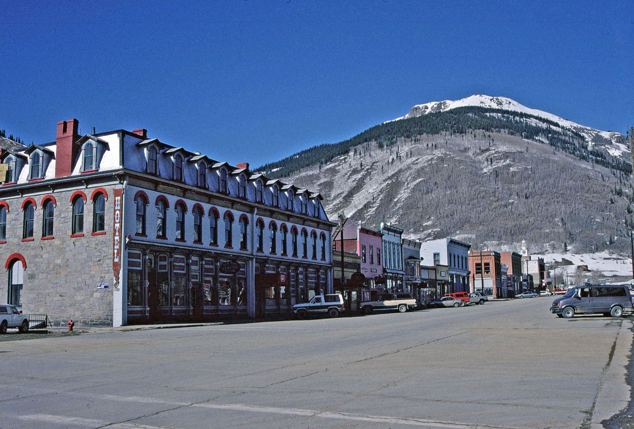 A view of the district with many buildings and a mountain in the background and snow in the foreground. The building in front has red trim and a mansard roof with dormers coming out.