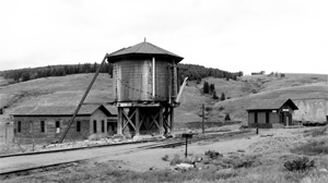 A black and white photo of the tracks with a water tank in the center and buildings on either side.