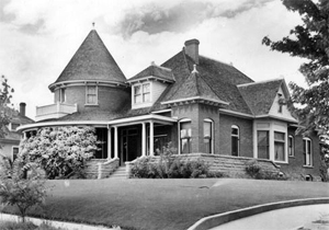 A black and white photo of the house with conical roof over rounded room and porch and multiple hipped and gabled roofs. A large yard and some trees stand in the foreground.