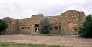 A view of the brick school with with entrance in the center and two wings on either side with some trees and a dirt field in front.
