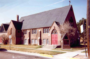 A view of the church from a mostly side angle with tall gabled roof, and cross gabled entrances at either end with brick walls. 