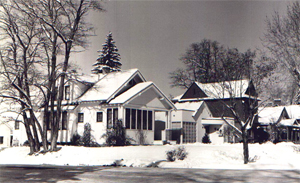 A black and white photo of some houses in the district with snow on the ground, large leafless and pine trees stand around the neighborhood.