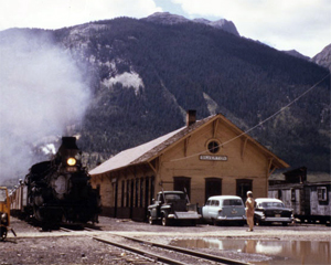 A picture of a train on some tracks next to a building and mountains rising in the background.