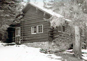 A black and white photo of the station with large log walls, door on the left and gabled roof. Around the site are trees and snow on the ground in front.