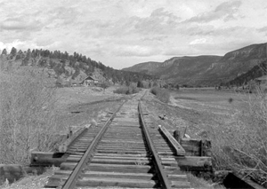 A black and white view of the railroad with tracks emerging from the perspective of the photographer and going out into the distance with hills and grass on either side.
