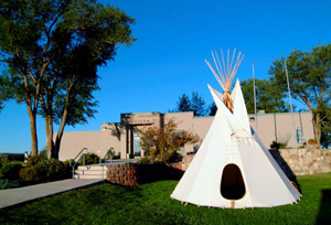 A photo of the site with tepee on the right in front of a wall with entrance on the left and large tree beyond that.  