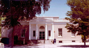 A view of the post office from the front with pediment supported by four columns and continuing walls on either side with two large trees on either side of the photograph, partially obscuring the building.