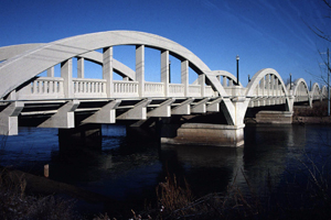 A view of the bridge from the side with large concrete arches over the bridge between abutments and railing along the sides.