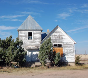 A photo of the church with fading white paint on a gable roof next to a tower with pointed hip roof and behind some short trees.