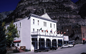 A view of a white building from an angle with mountain behind it. The building has a covered balcony with wooden pillars supporting it. 