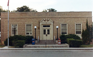 A photo of the post office from the front with light colored walls, white entrance in the center and two vertical windows on either side.