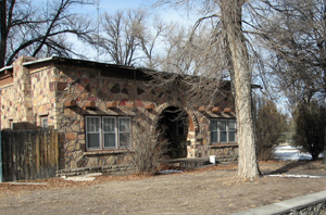 A view of a building with flat roof and large arched entrance between two sets of three windows. There is a large leafless tree on the right and trees behind.