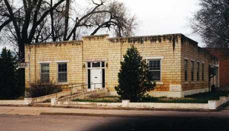 A view of the building with light brick walls and vertical windows. There is a door in the center of the building with railed path leading to the street out front.