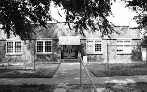 A black and white photo of th school with walkway leading up to the main entrance and awning in front. On either side are two large windows and above are leafy branches hanging down. 