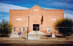 A view of the school from the front with red brick walls, small curved gable on top and above the central entrance below with chain linked fence before the building.