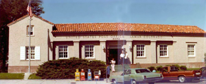 A picture of the post office with red pitched roof and gabled section on the left over sand colored walls with pilasters in the center.