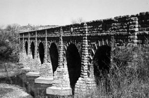 A black and white photo of the stone brick bridge with several large abutments and river on the left.