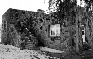 A black and white photo of a stone building in the park with stairs leading up to the left and window on the right with door just visible on the right wall.