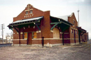 A picture of the building with red brick walls, arched window and gables on the top and sides with overhanging green roof. 