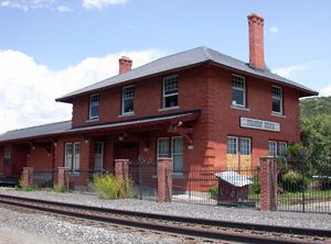 A view of the depot at an angle from across the railroad tracks. The depot has two stories with hipped roof and two chimneys on either side, on the left is an overhanging pitched roof section. 