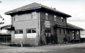 A photo of the depot in black and white with two stories and hipped roof over pitched roof on the right side. 