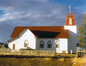 A view of the white church from the side with red pitched roof and cross gabled section on the left and tower with cross on top on the right.