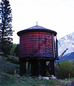 A picture of the tank with red sides and conical roof with foliage and mountains in the background. 