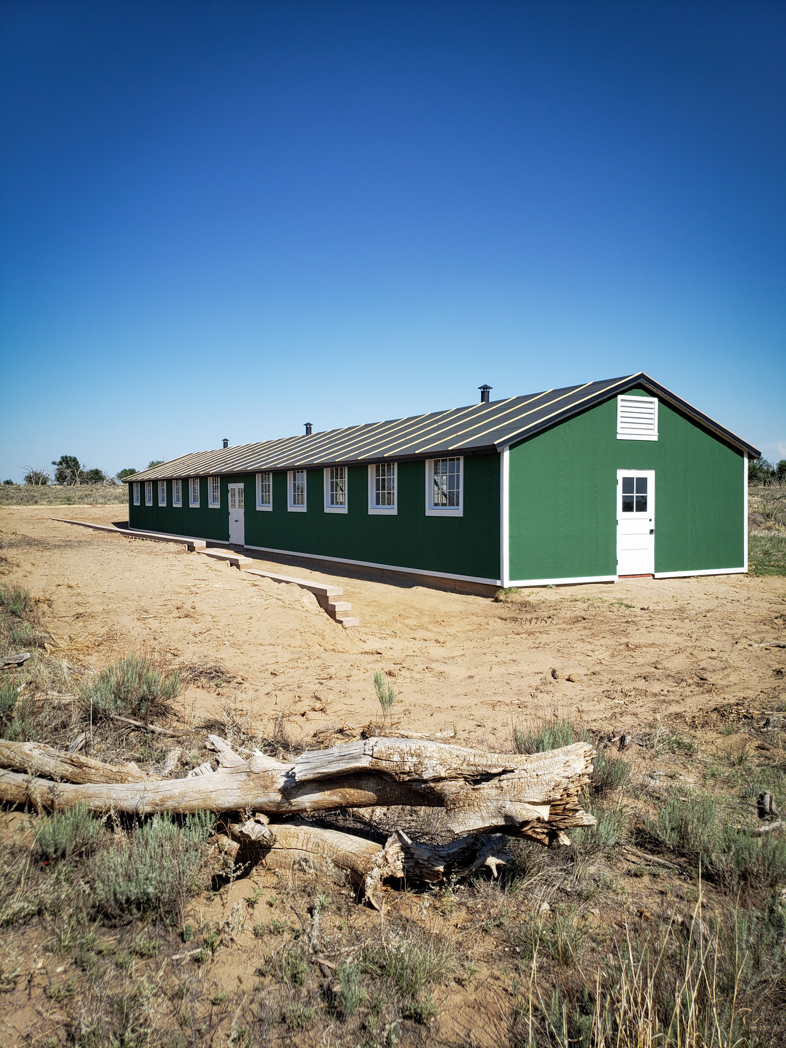 photo of the completely restored Amache camp recreation building in the distance. The long green building with white trim is contrasted against the flat arid land surrounding it.