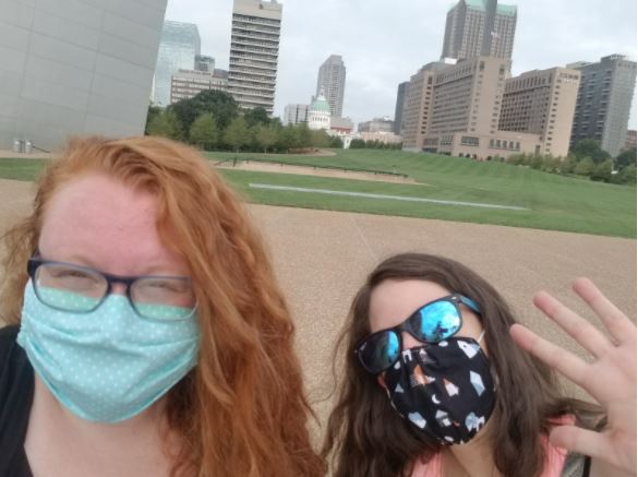 Photo of Amy Nilius and Kristin Chiesi, on a quick walk in St. Louis. They are taking a selfie, wearing sunglasses and face masks due to COVID-19. Behind them the high-rise buildings of downtown St. Louis are visible.