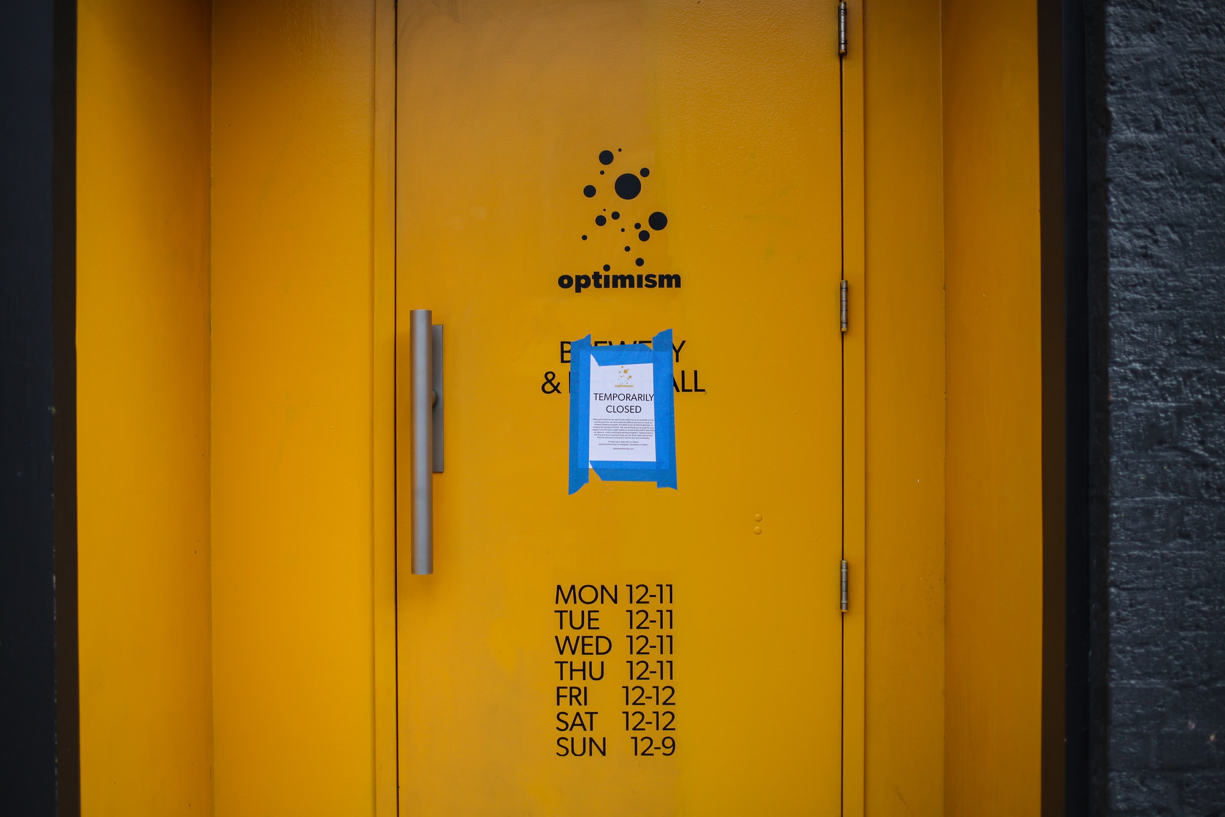 Photo of the bright yellow industrial front doors of a business which has been closed due to the pandemic of 2020. The business name appears to be Optimism, and there is a printed paper sign taped to the door beneath the name of the establishment, which says "Temporarily Closed." At the bottom of the door are the hours of operation for the business. 