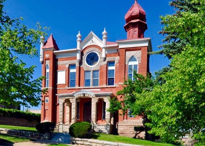 Photo of Temple Aaron, after the completion of a preservation project. The handsome building appears to be made of red sandstone brick and has white and blonde trim. There is a front porch made of blonde brick, and a large round window in the building directly above the door. There are gothic style arched windows and decorative finials along the roof of the building.