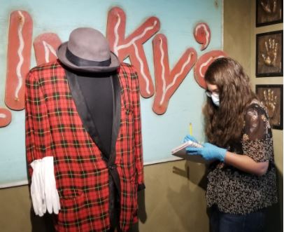 Photo of Kristin Chiesi, completing a condition report on Blinky the Clown's Jacket before taking it into History Colorado's collection. Kristin is seen here, standing in front of a mannequin on which Blinky's iconic red plaid jacket, black bowler hat, and white gloves are positioned. Kristin is wearing blue latex gloves and holds a clipboard. Because of COVID, she is also wearing a face mask.