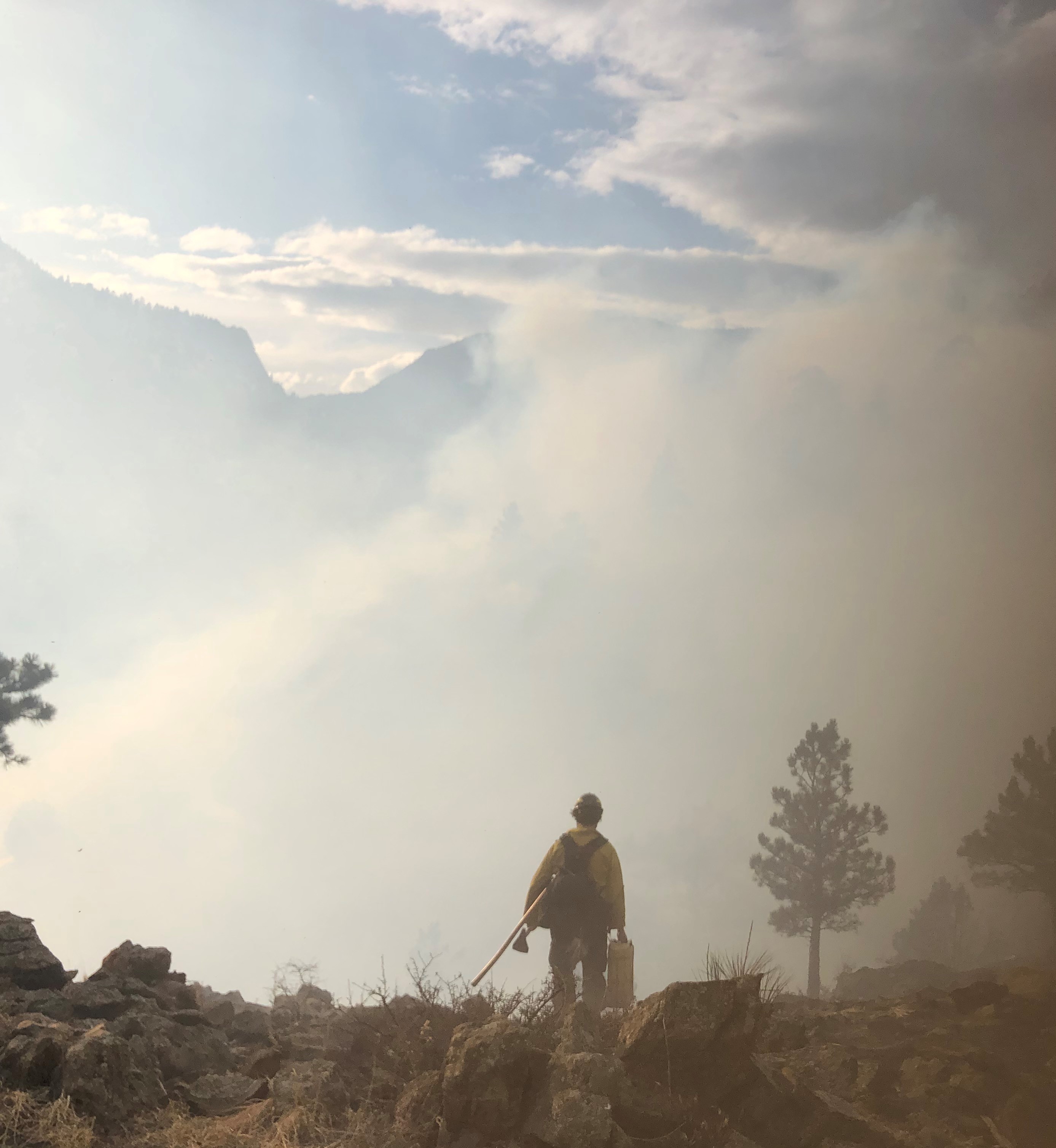 Photo of a wildfire burning in the valley. A firefighter stands at the rocky edge of the mountain, overlooking the burn site as a blanket of thick gray smoke covers the valley. Barely visible through the smoke are the mountain tops across the valley.