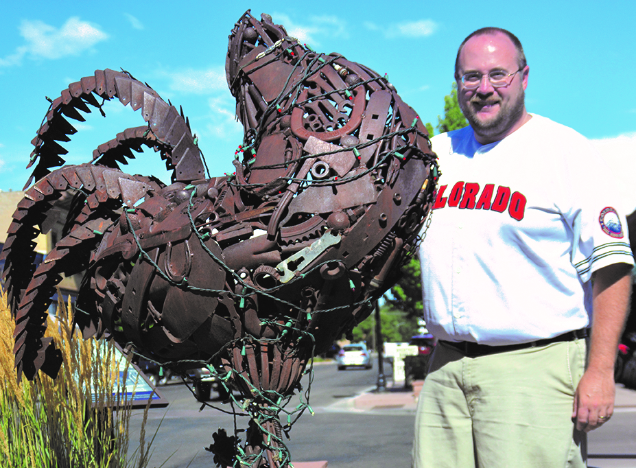Photo of author Derek R. Everett, standing next to a sculpture of Mike the Headless Chicken, located in the town of Fruita, Colorado. The sculpture is made of reclaimed metal objects, such as a horseshoe, an axe, bolts, etcetera. It is a sunny day, with blue skies overhead, and the author is smiling, wearing a white Colorado baseball jersey with red lettering, and khaki pants.