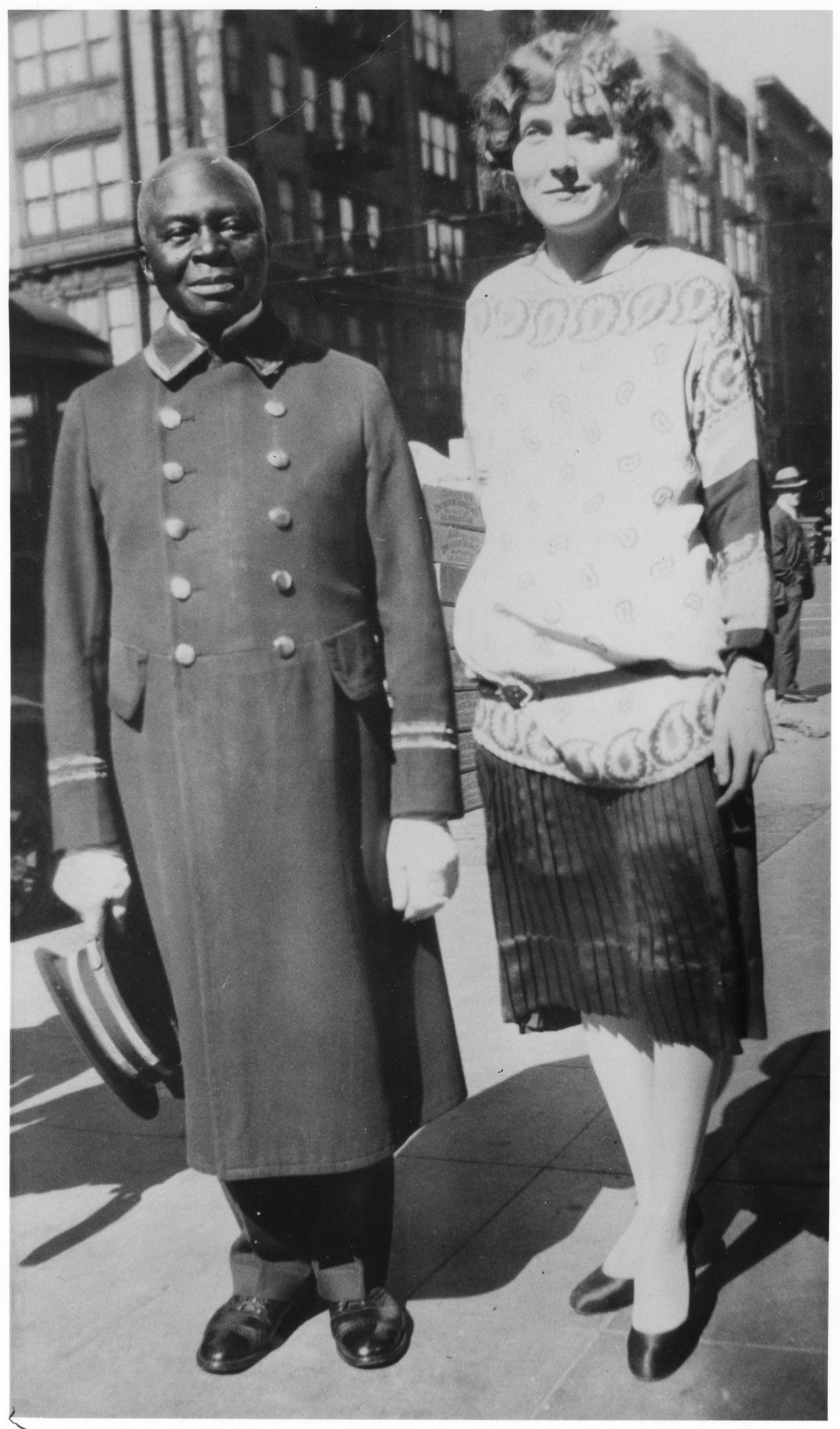 Photo of Frank Loper, circa 1925, when he was working as a doorman for the Antlers Hotel. He is pictured with Mrs. Glen Hutchinson, the hotel's bookkeeper. He is wearing a long doorman's coat with two stripes near the cuff of each the sleeve and ten shiny metal buttons on the front of the collared jacket. In his right hand, he is holding his uniform hat. Mrs. Hutchinson wears a skirt, tights, flat black shoes, and a lacy long sweater. They are standing in the sidewalk, presumably in front of the hotel.