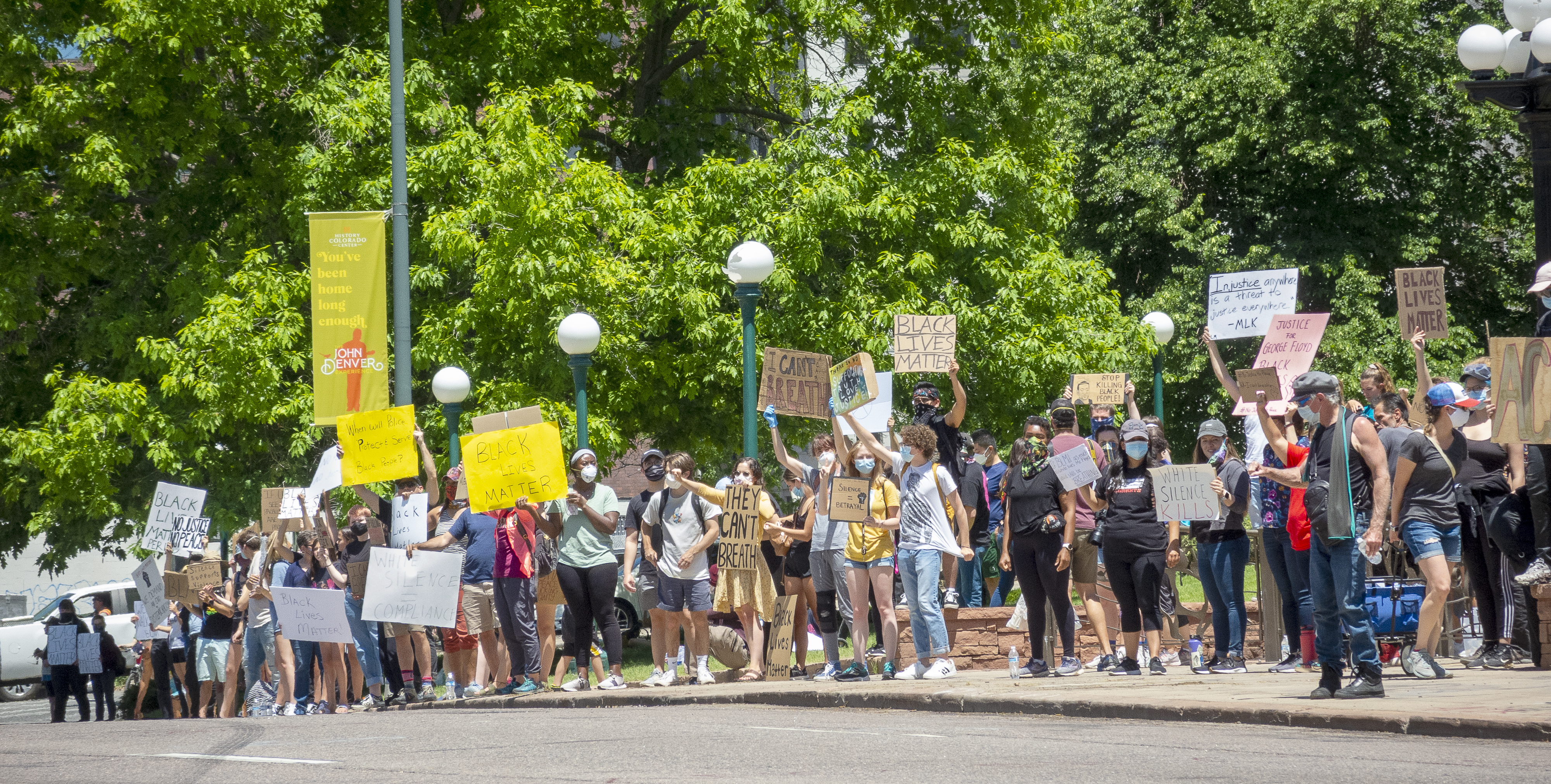 Photo of  Black Lives Matter protestors in Denver in 2020. A large group of people stand along the sidewalk, holding up signs and wearing face masks due to Covid-19. Many of the signs read "Black Lives Matter" and other say "I Can't Breathe" and "No Justice No Peace."
