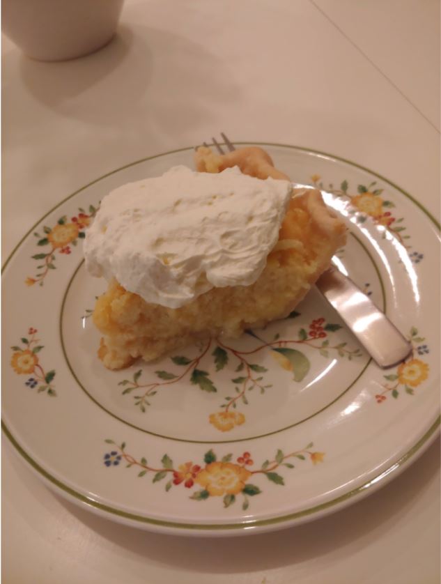 Photo of a piece of lemon buttermilk pie, ready to be eaten. It sits on a china plate adorned with brightly colored flowers, and a fork sits at the ready next to the slice of pie. The plate is on a table covered with a light-colored table cloth, and the bottom half of a white china cup is barely visible in the corner of the photo.
