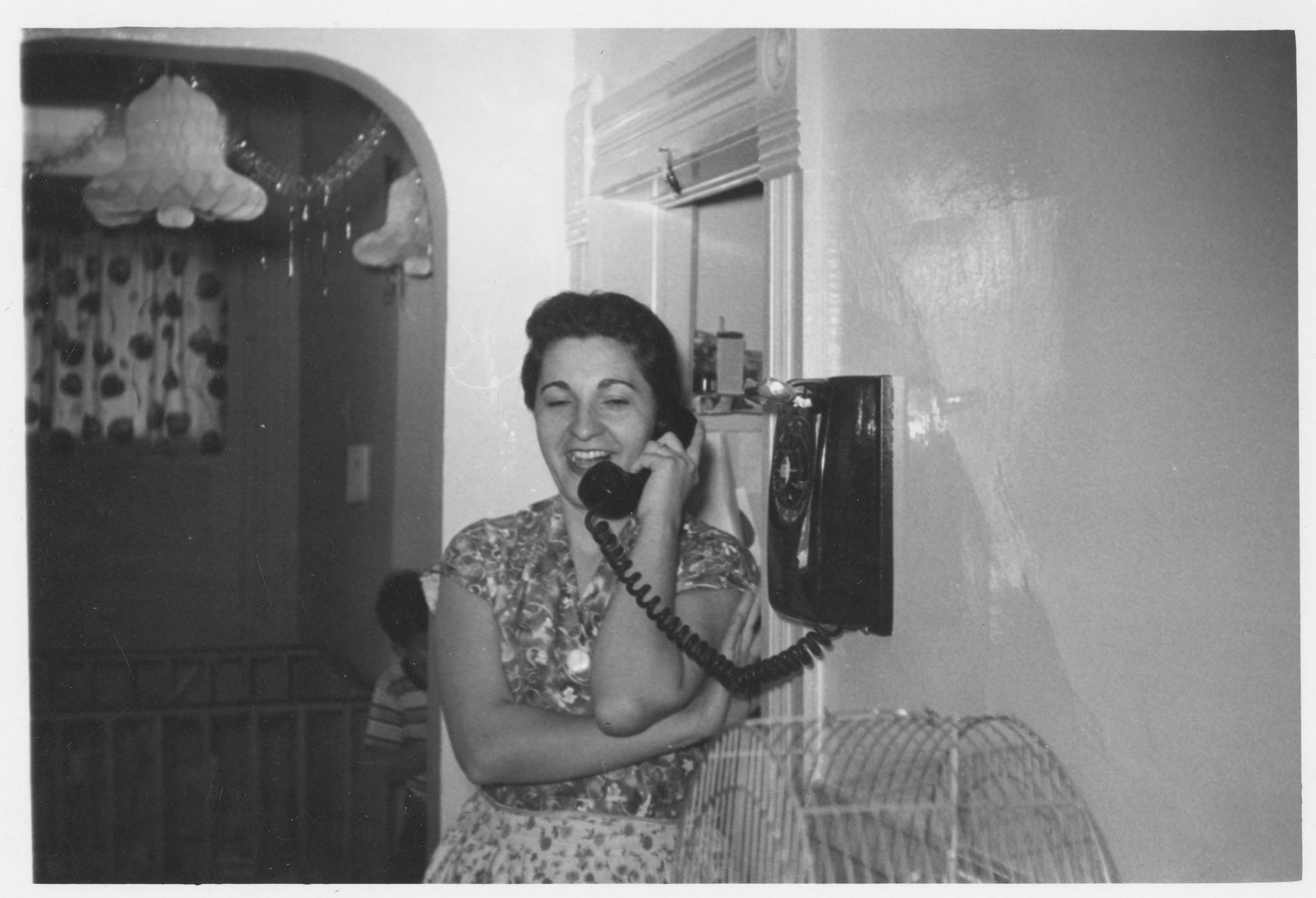 Photo of a woman talking on the telephone, around 1955. She is wearing a dress with a belt, and she has short dark hair. She is standing in a hallway, leaning against the wall to talk on the wall-mounted telephone. A holiday decoration made of paper in the shape of a bell hangs from the ceiling behind her. She is smiling while she chats on the phone.
