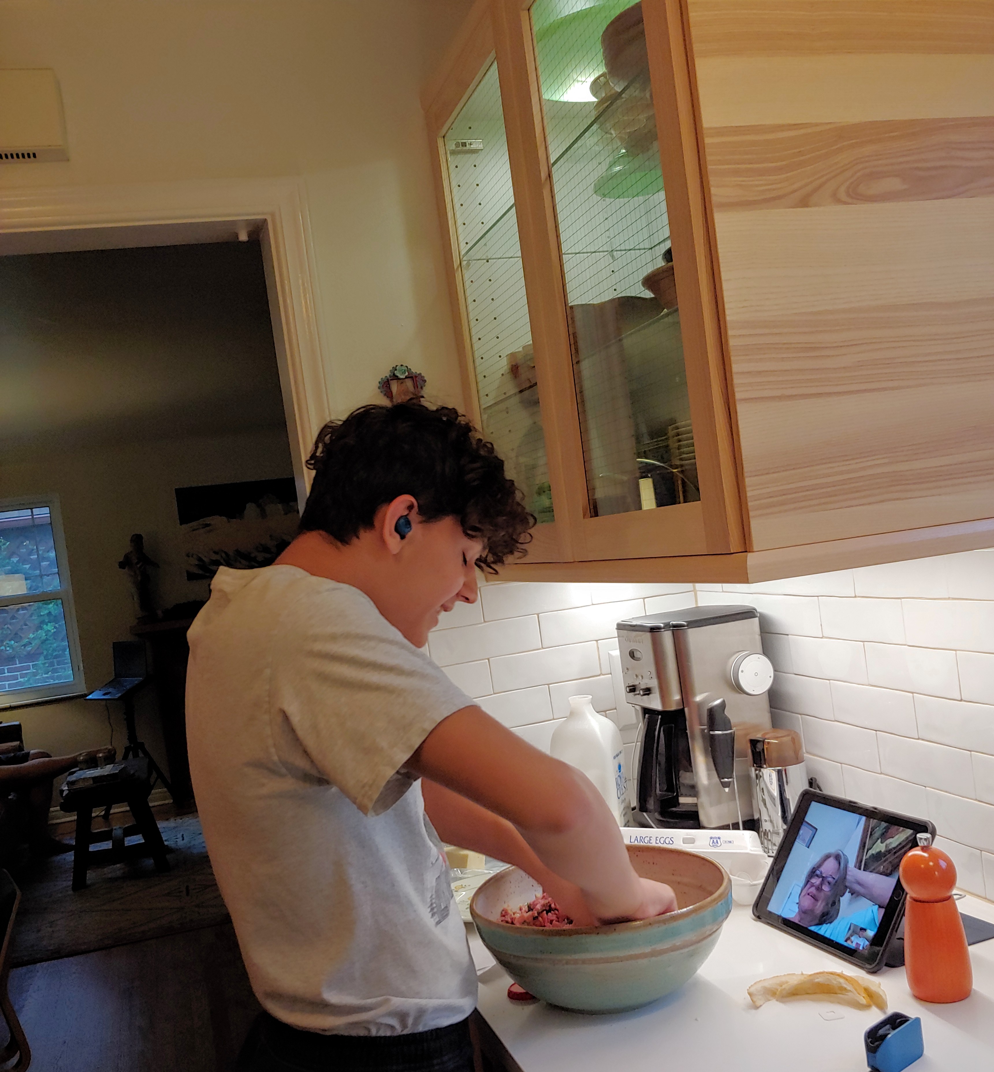 Photo of a young man, looking at his computer tablet and learning to make meatballs with his grandmother over virtual meeting. He is mixing ingredients in a large bowl with his hands, while his grandmother is seen on the computer screen, giving him instructions.