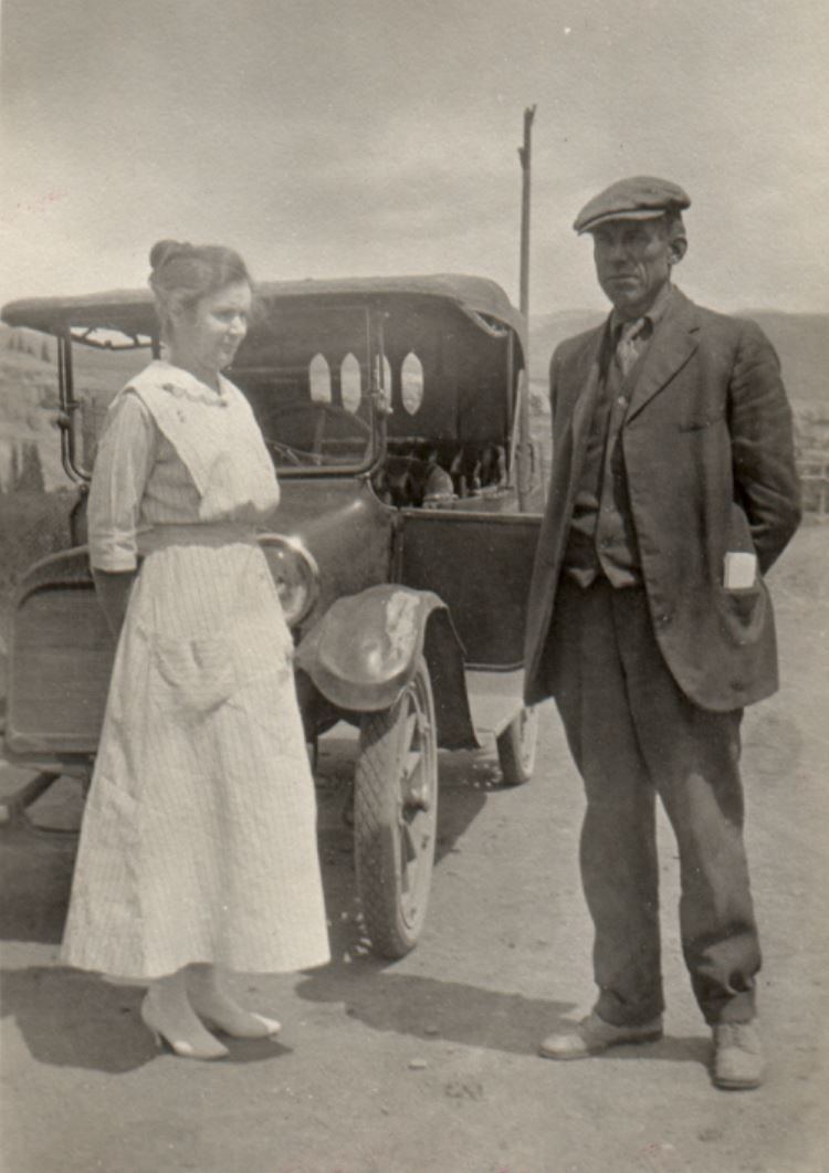 Photo of a couple in the early 1900s, standing in the road next to their first car, a 1917 Dodge. The driver's side door is open, and the couple are looking ahead in different directions. The woman is wearing a dress with white boots, her hair up in a bun at the back of her head. The man is wearing trousers, leather shoes, a shirt, tie, and waistcoat, with a jacket. He has a flat driving cap on his head, and folded papers in his left jacket pocket.