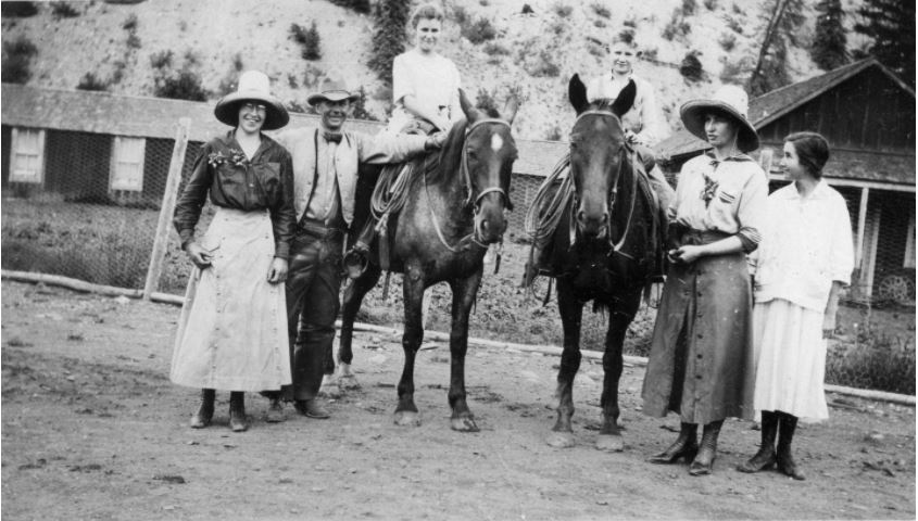 Photo of a group of people out for a walk and horseback ride. There are two horses, atop one of which there is a young woman ready to ride. A man and three other women stand beside the horses, dressed in wide-brimmed hats and riding attire for the  period.