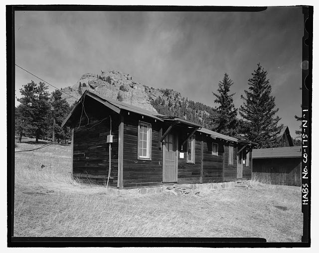 Photo of small twin cabins, side by side on the ranch. They are wooden framed cabins with wood horizontal siding, unlike the other ranch buildings which are log cabins. These cabins were residence buildings for female worker, and feature a door in the center of the cabin front, flanked by long 8-paned windows on either side.