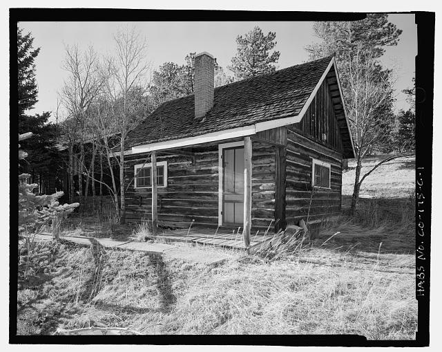 Photo of small one-story log cabin with a front door on the right side of the front of the building, and a small 8-paned window on the left side. The roof is pointed and a slender chimney comes out of the middle of the roof.